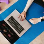 Top view of crop female in activewear sitting on yoga mat on floor and using laptop